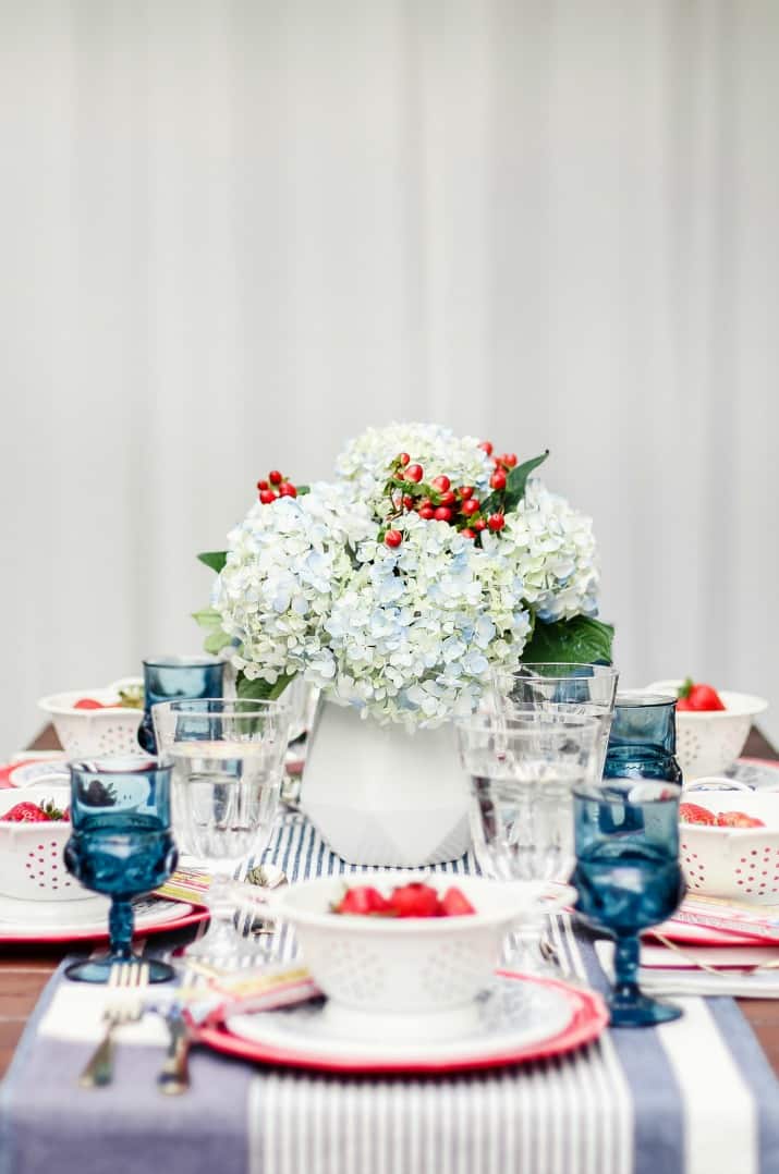 4th of July centerpiece of blue hydrangeas in white vase sitting on outdoor dining table.