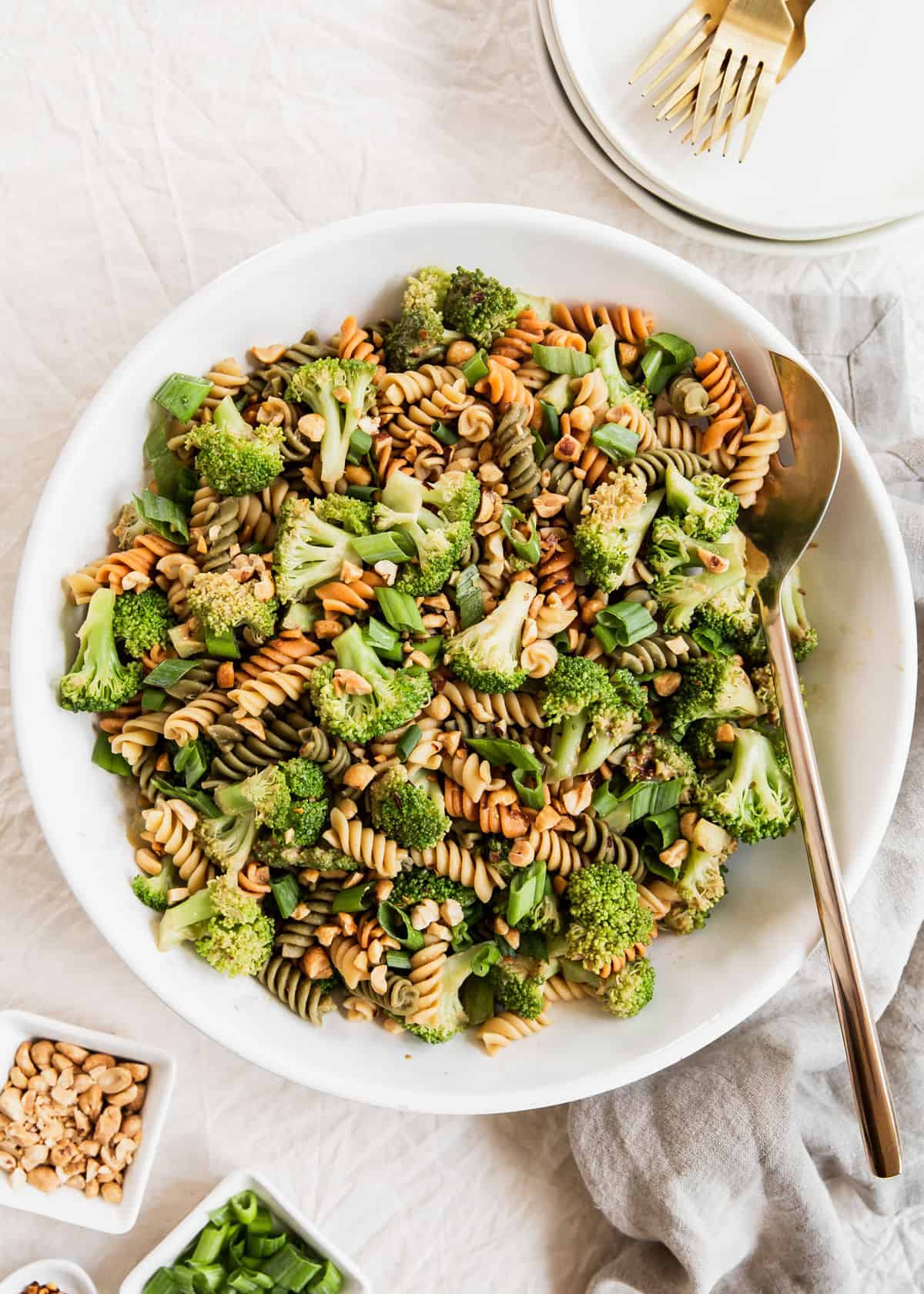 large white bowl with broccoli and pasta salad, large serving spoon, and surrounded by plates and garnish bowls.