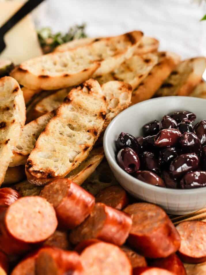crostini with grill marks next to smoked sausage cubes and black olives.