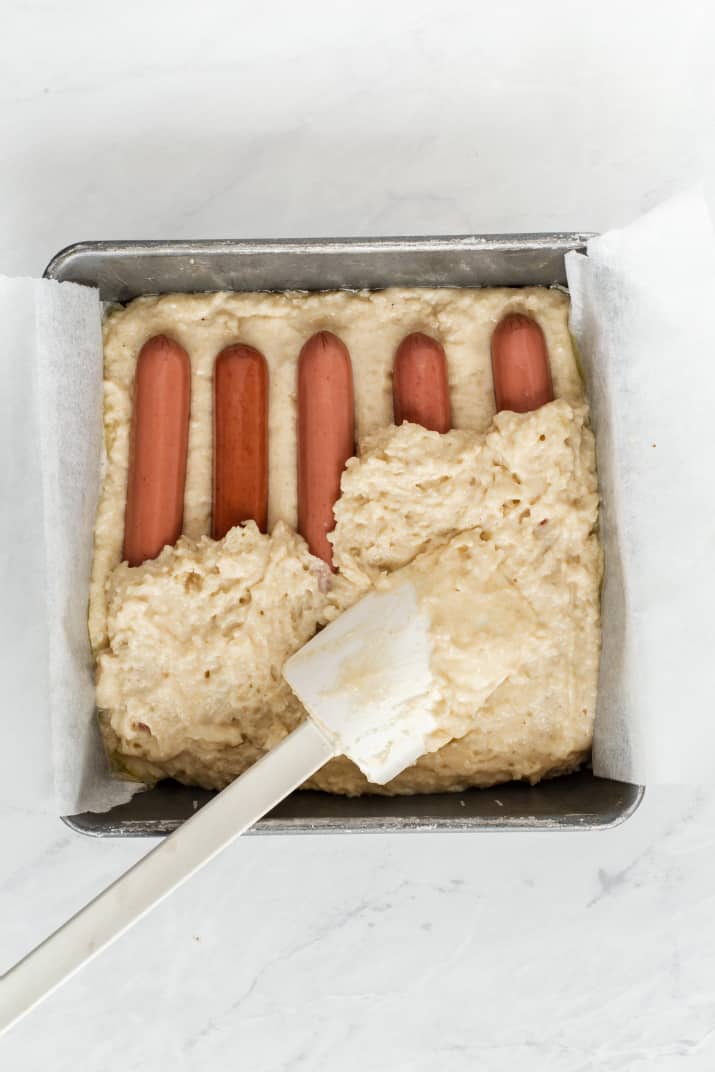 batter in square baking pan with hot dogs lined up inside.