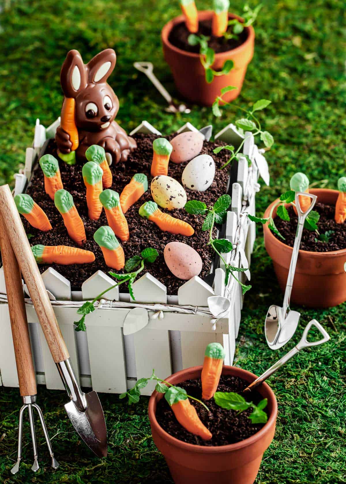 dirt cake in white fenced garden container, decorated with chocolate bunny, candy carrots, and chocolate eggs.