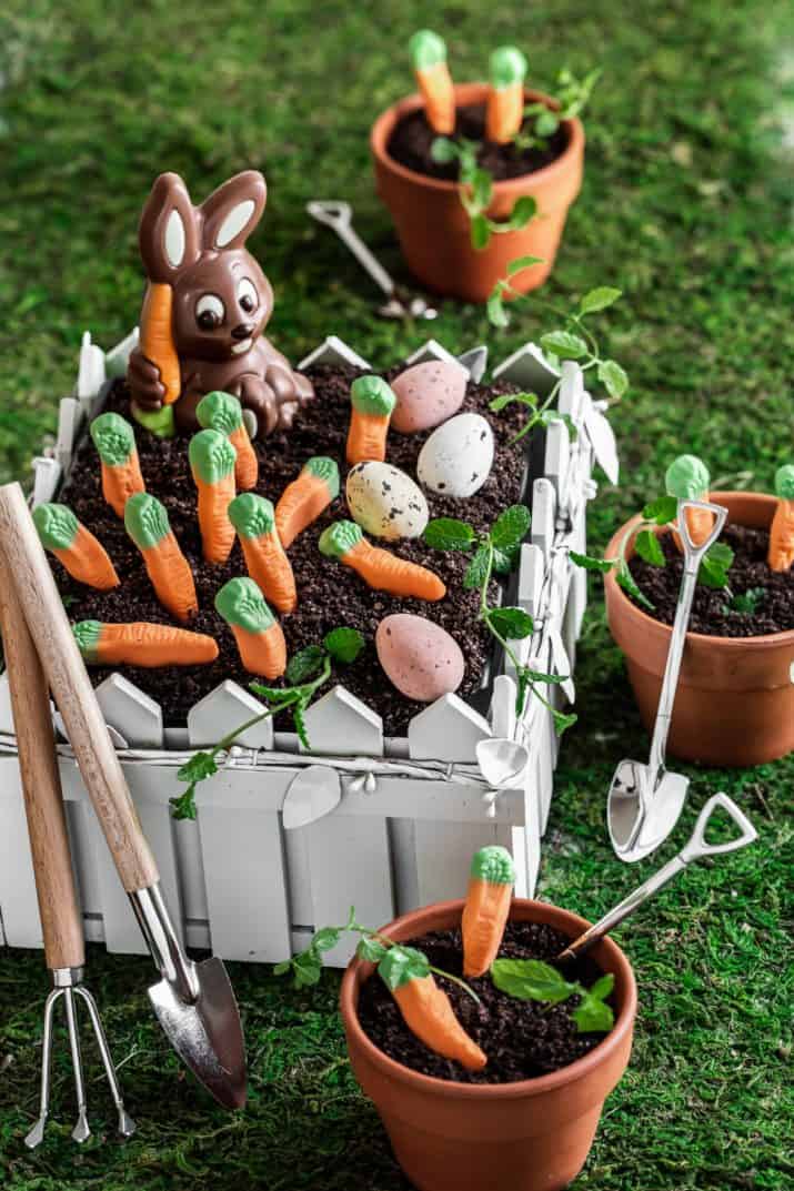 dirt cake in white fenced garden container, decorated with chocolate bunny, candy carrots, and chocolate eggs.