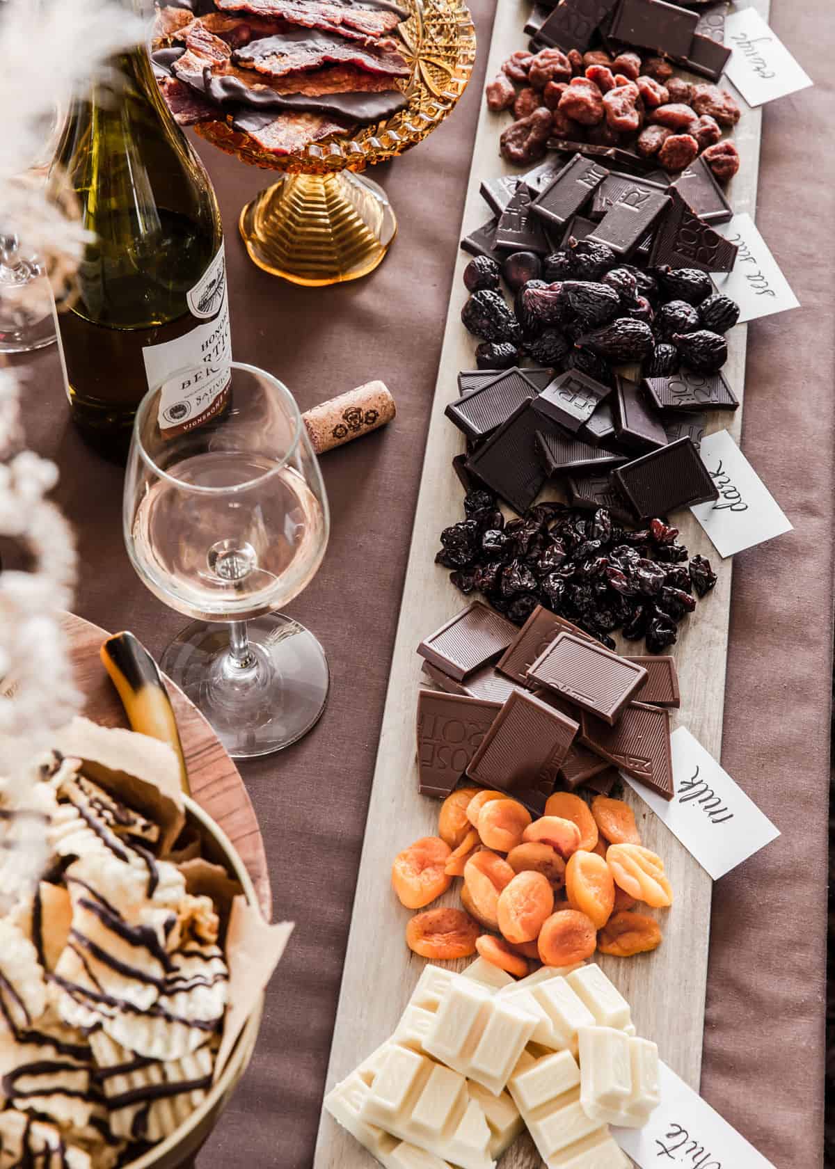 long wood plank filled with chocolate squares and dried fruit on brown tablecloth.