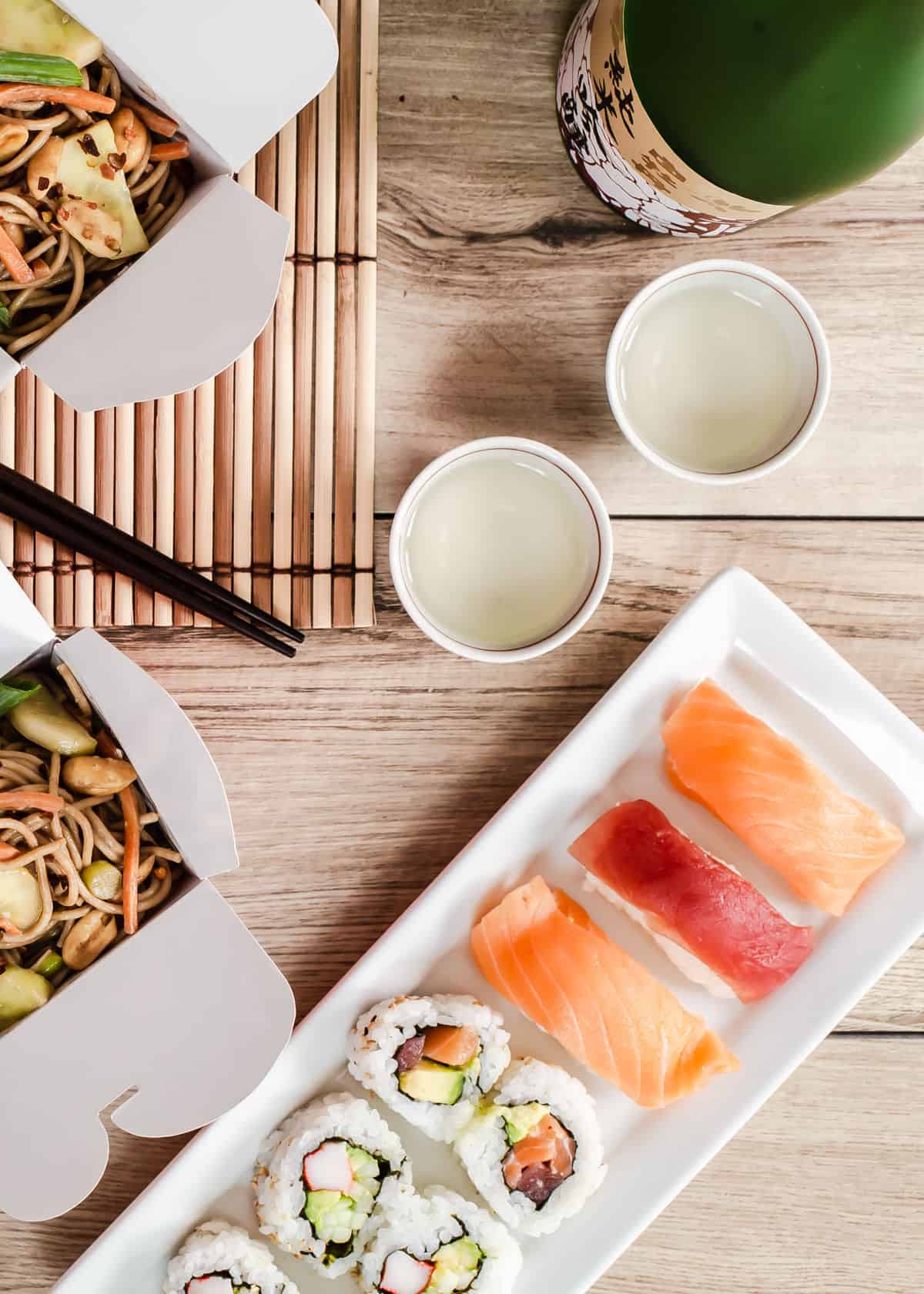 overhead view of sushi on long white dish surrounded by sake cups and Chinese takeout boxes.