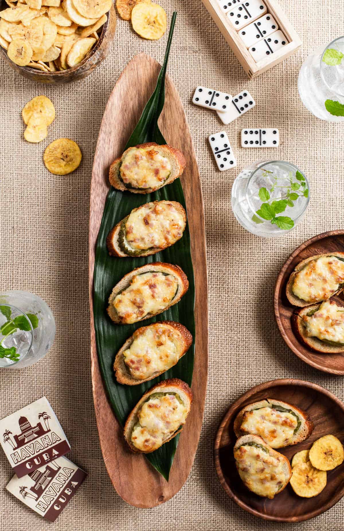 party canapes on wood tray with burlap tablecloth, surrounded by dominos, drinks, and plantain chips.