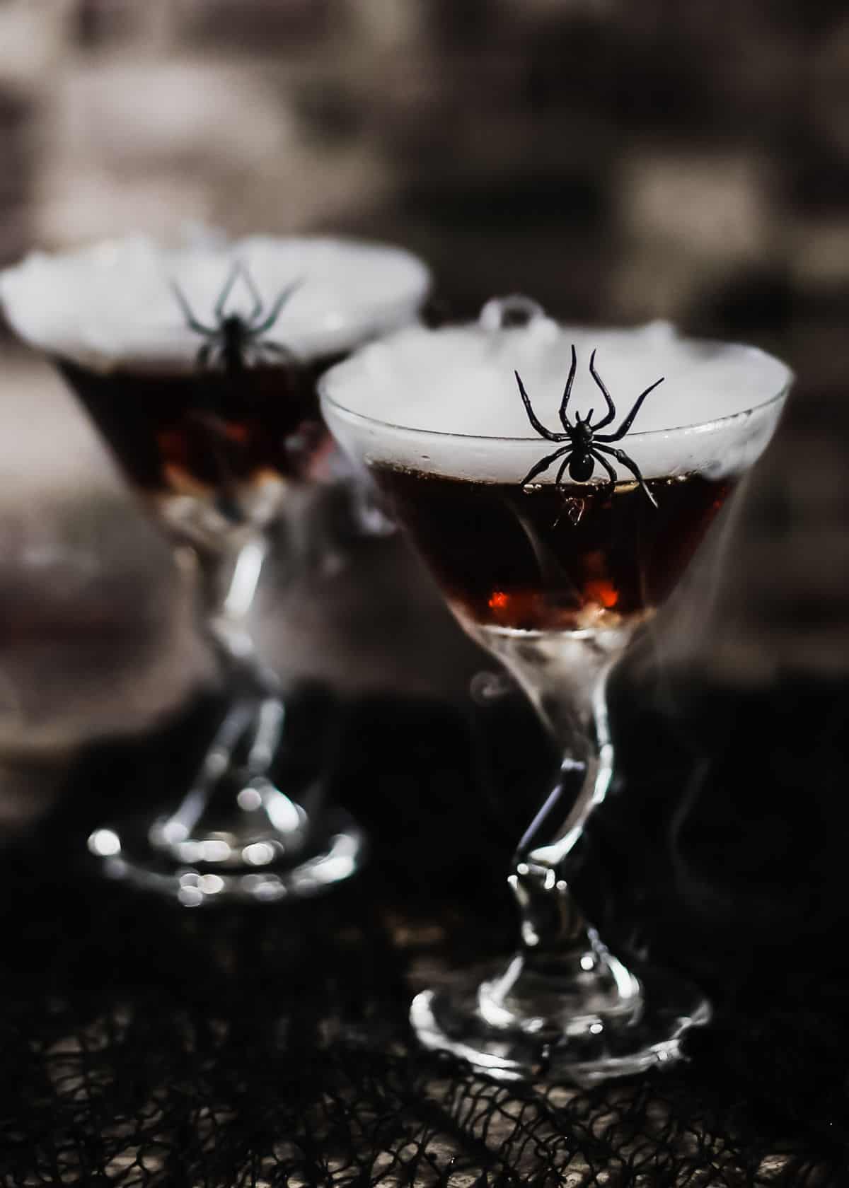 dark cocktail in martini glass with dry ice smoke and spider on side of glass.