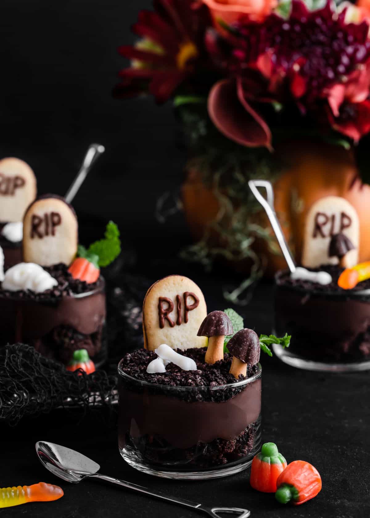 Halloween graveyard dessert cups on black table with flowers in background.