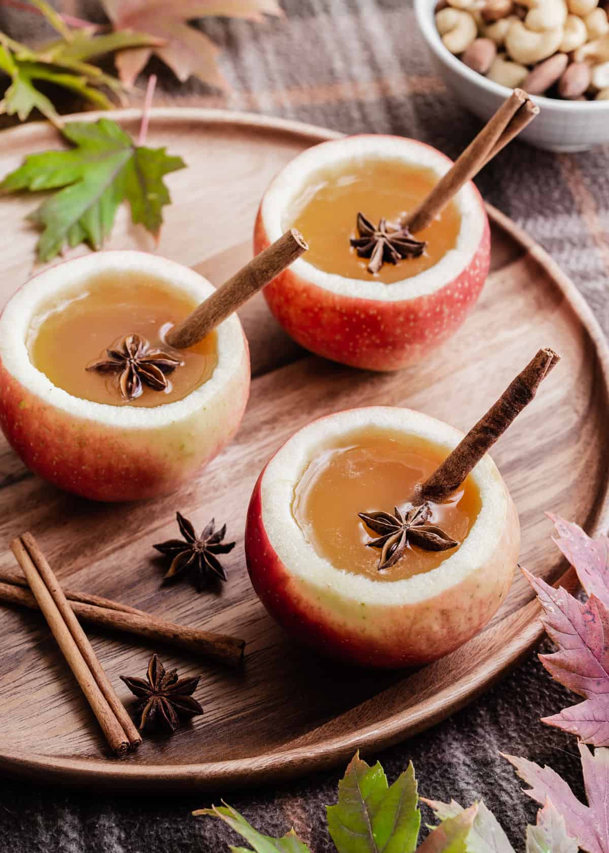 three hollowed out apples on wood tray, with cider drink served inside, garnished with cinnamon stick and star anise.