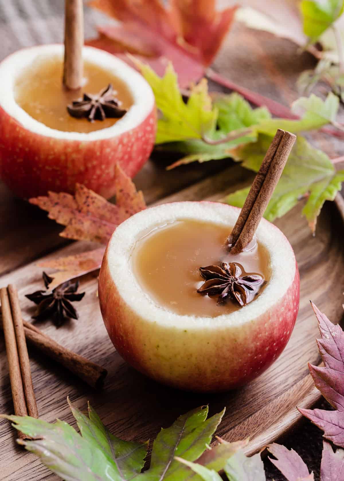 apple cider in apple cups, with cinnamon stick garnish, surrounded by fall leaves.