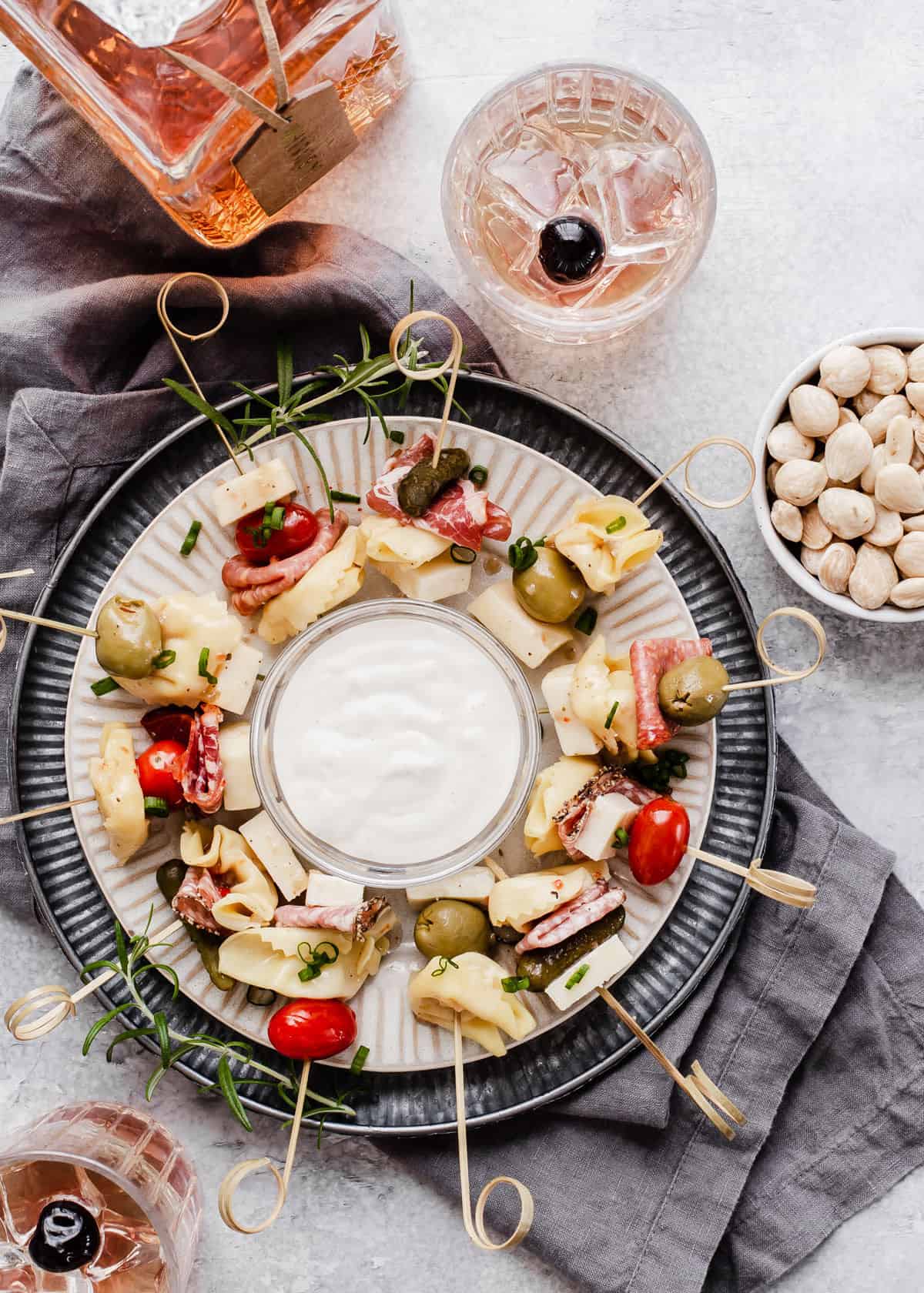 round dish of skewered appetizers with tortellini, cured meats, pickles, tomatoes and olives, surrounded by drinks.