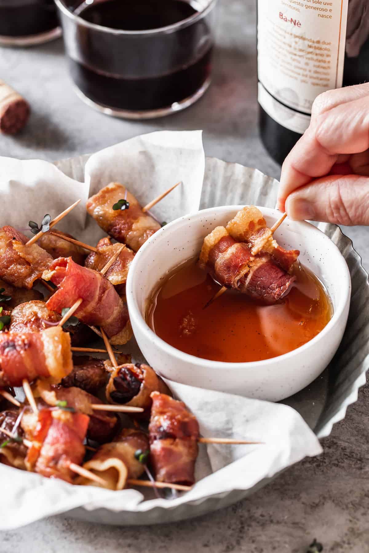 round dish holding bacon wrapped appetizers with hand dipping one into dipping sauce.
