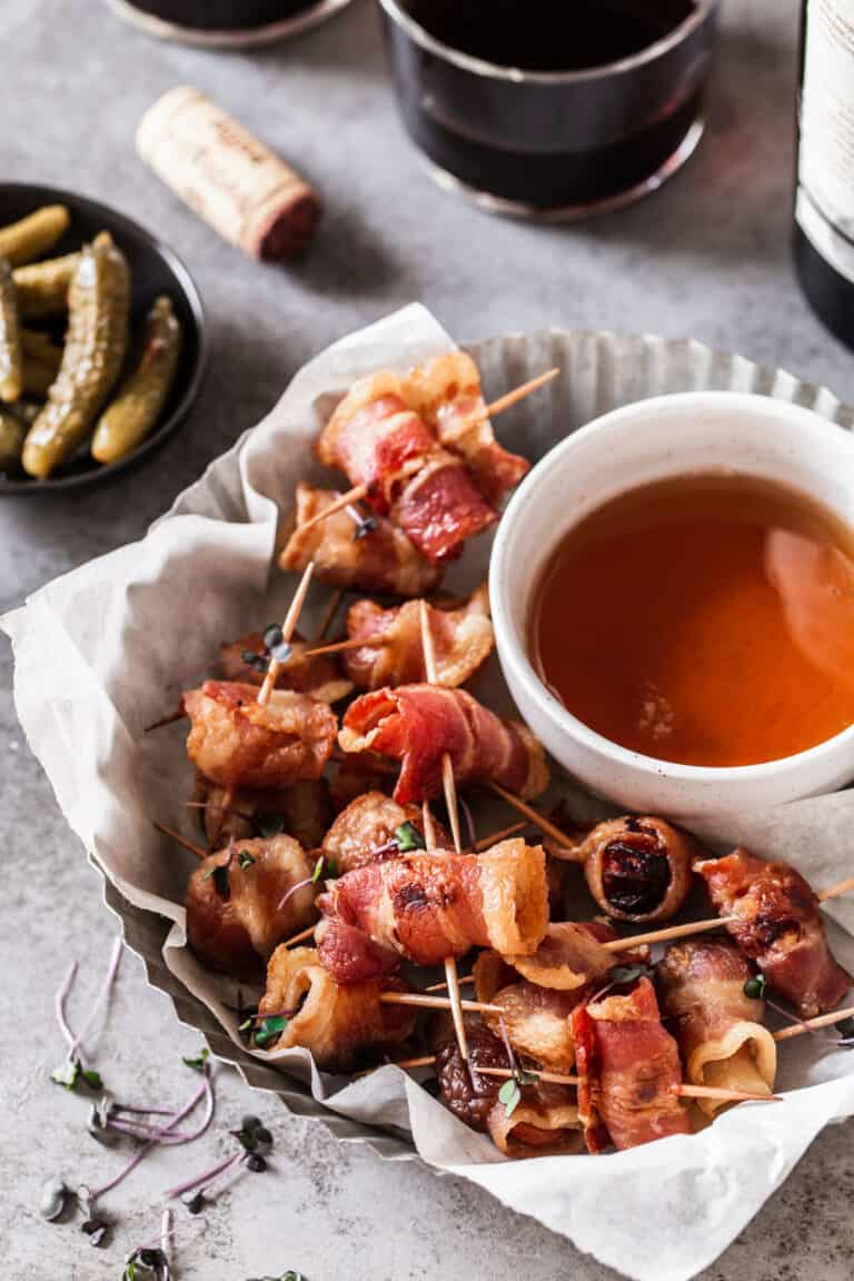 round silver dish filled with bacon wrapped appetizers with small white dipping sauce bowl, surrounded by wine and baby pickles.