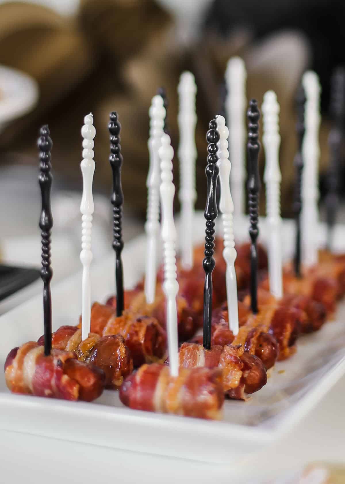 little smokies wrapped in bacon on white tray with black and white picks inserted.