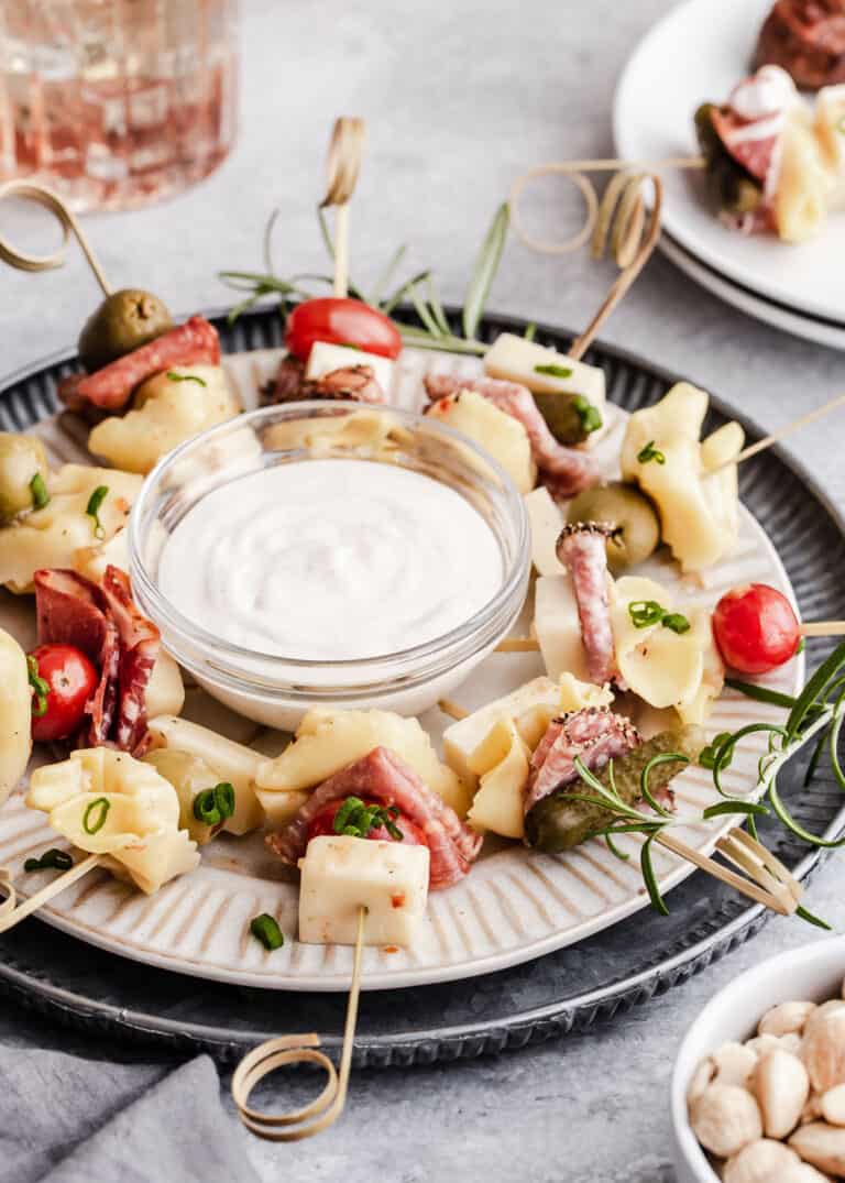 round dish of antipasto skewers with tortellini, meats, cherry tomatoes, olives and pickles, with white dip bowl in center.