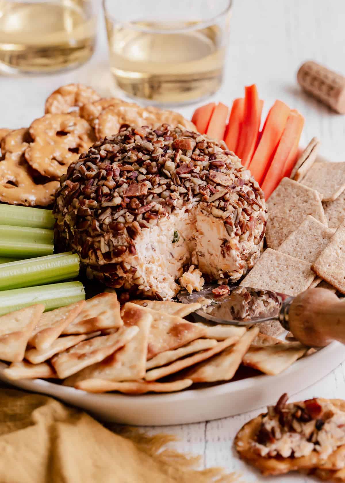 cheese ball covered in chopped pecans and bacon bits, surrounded by crackers, carrot sticks and celery sticks.