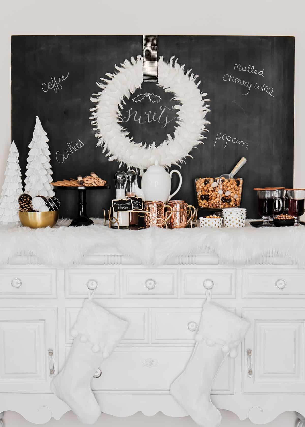 white buffet table with desserts and drinks with chalkboard sign and white wreath on wall.