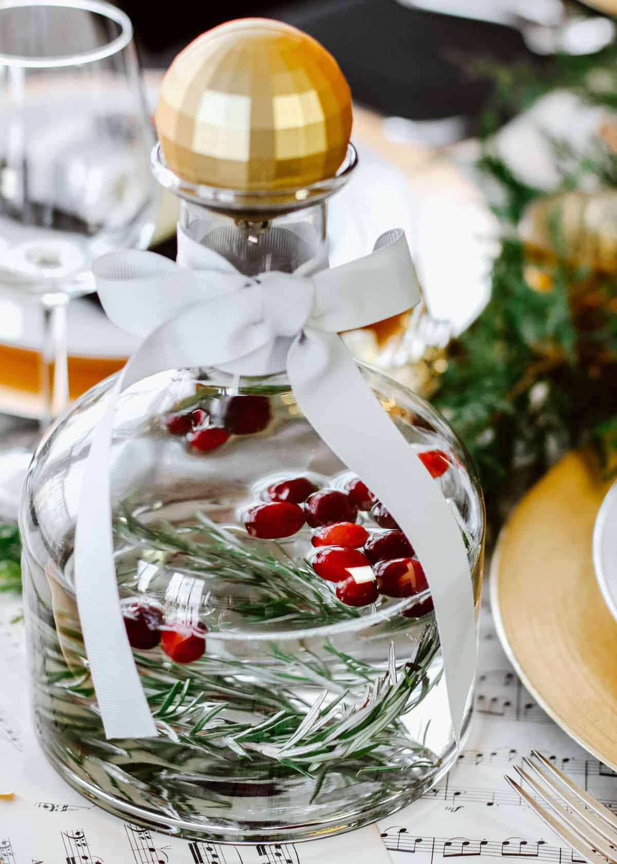 carafe of water with rosemary and cranberries inside, on table with ornament bottle topper.