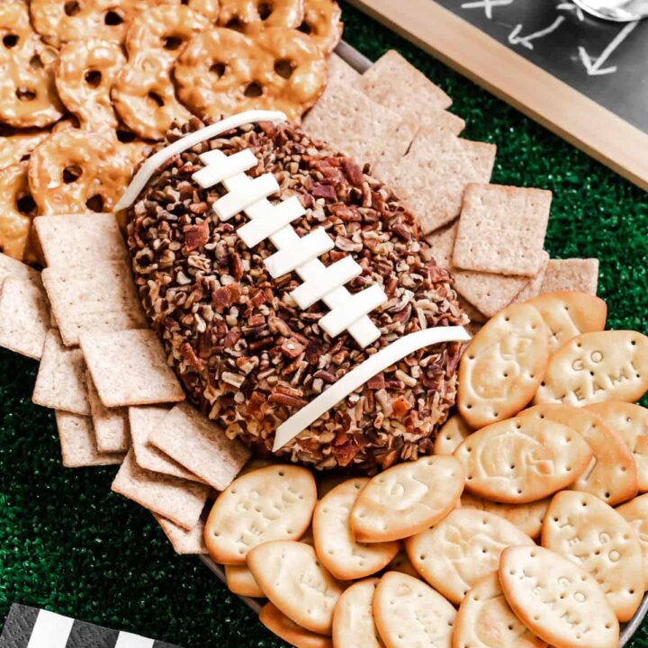 football shaped cheese ball on oval platter surrounded by crackers and pretzel crisps, on green turf grass with football party decorations.