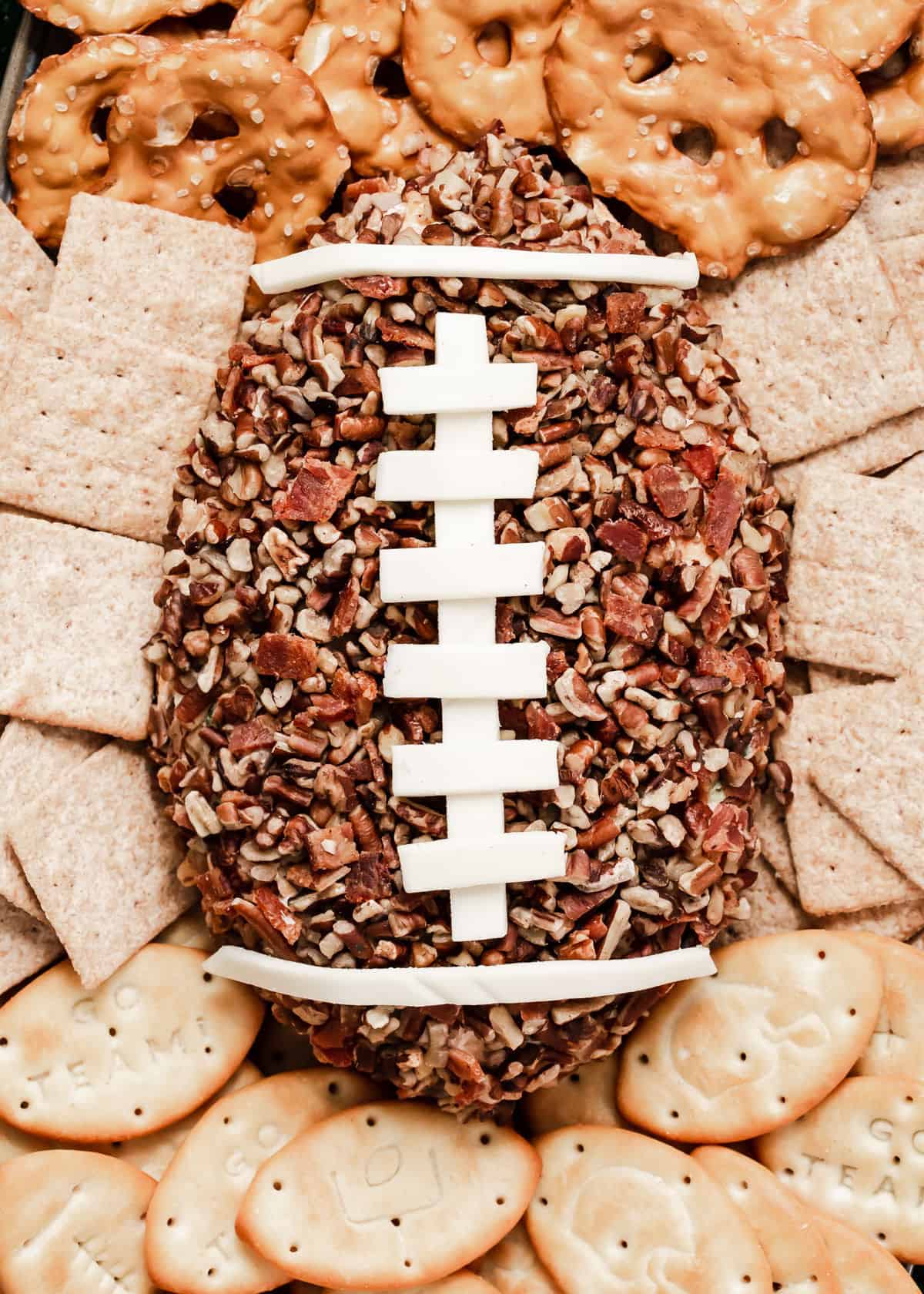 close up of cheese ball shaped like a football covered in chopped pecans and bacon bits with white cheese stitching.