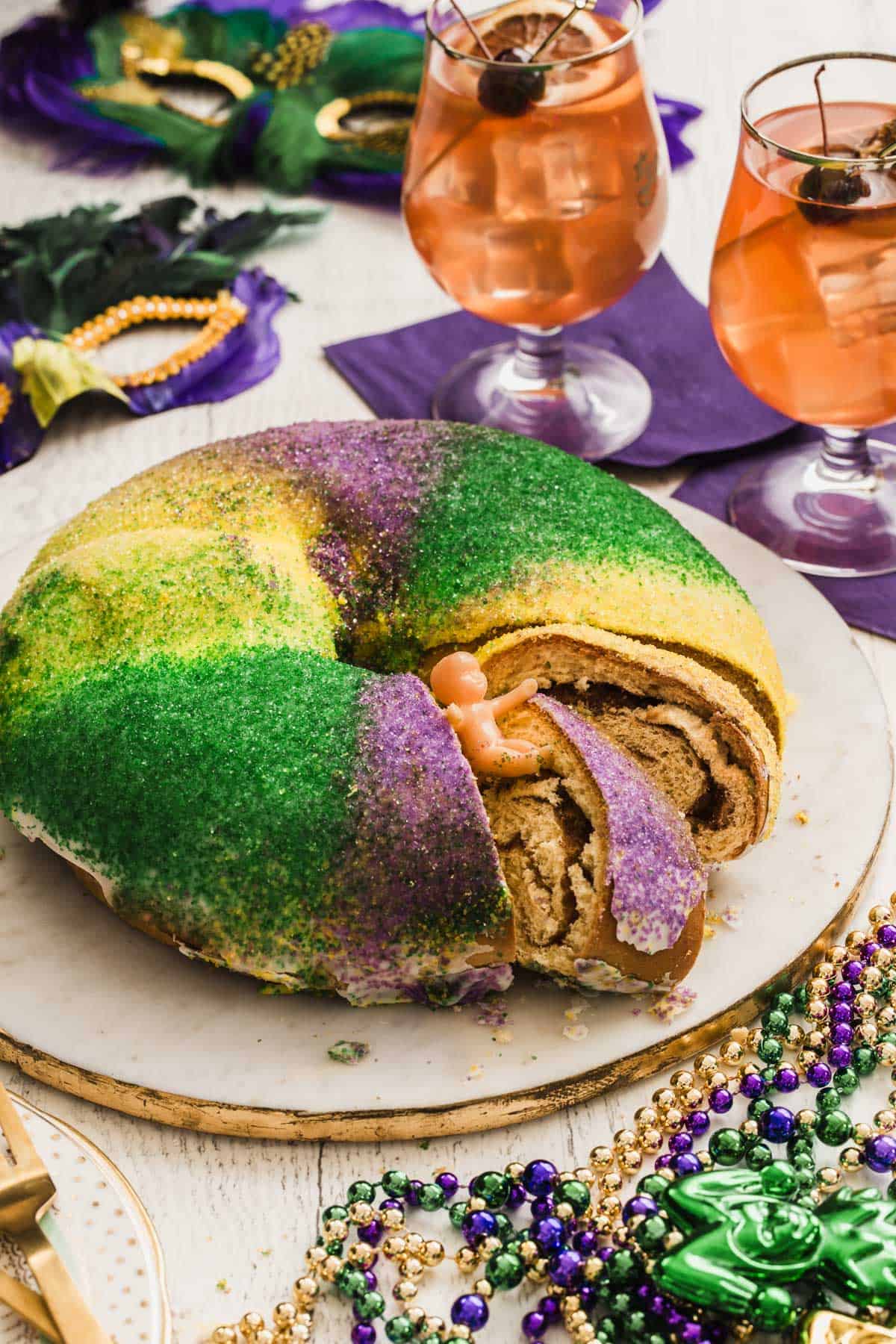 Mardi Gras King cake on white platter surrounded by mask, beads, and hurricane drinks.