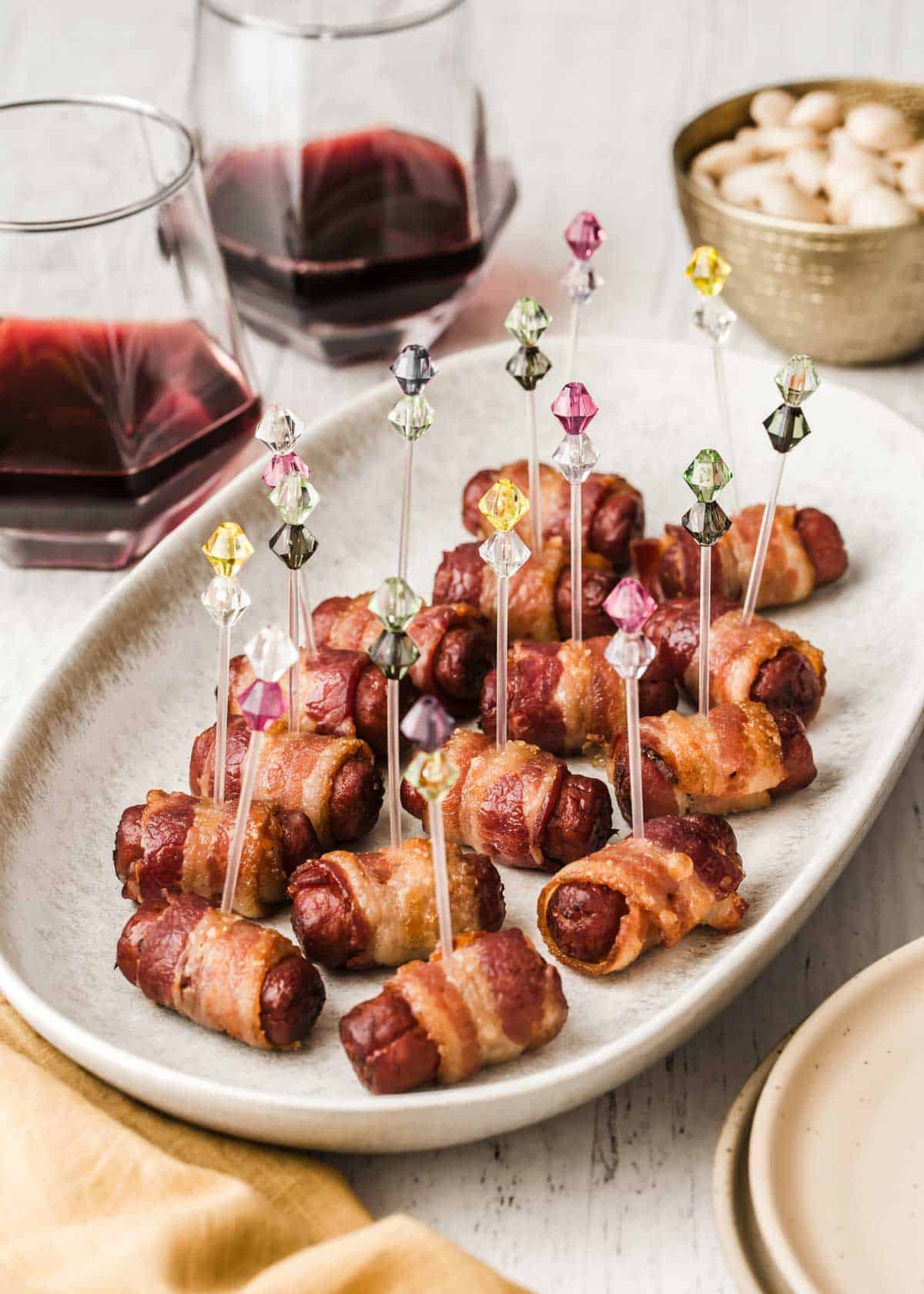 bacon wrapped little smokies on cream platter with red wine and bowl of nuts in background.