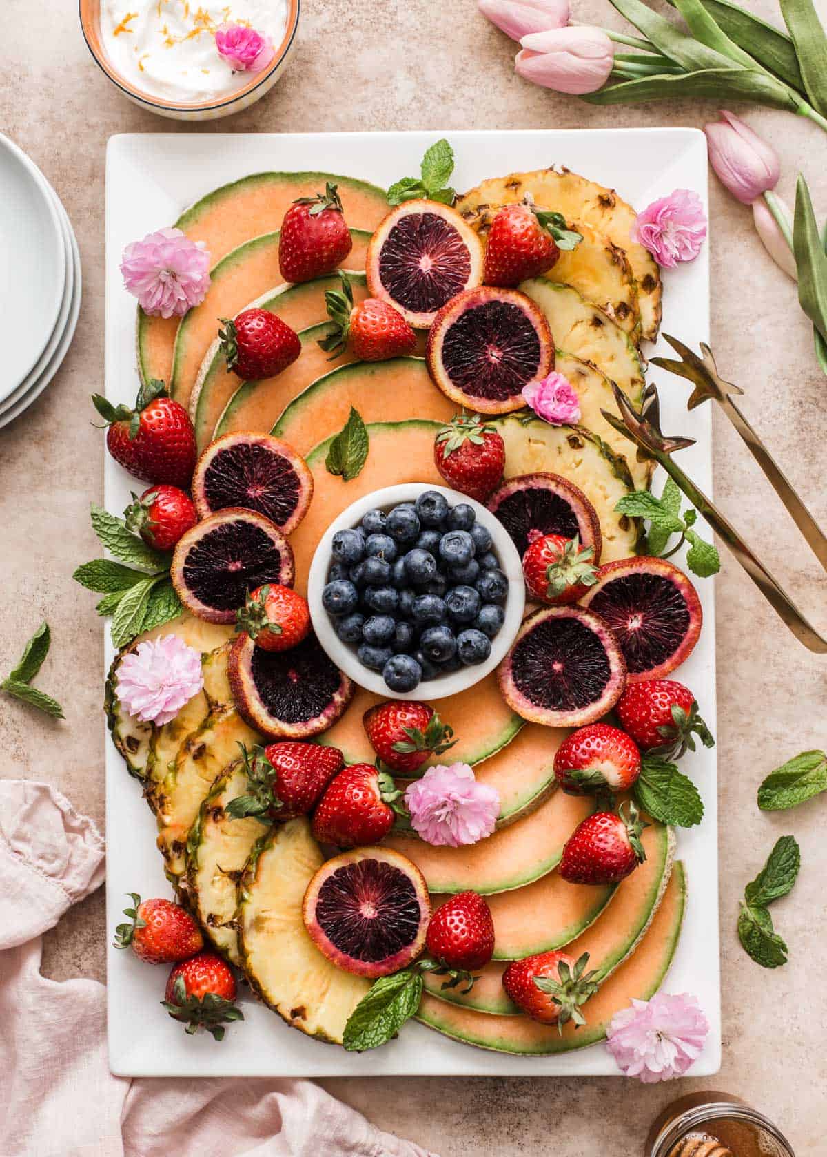 large white platter with sliced cantaloupe, pineapple, blood oranges, whole strawberries and blueberries.