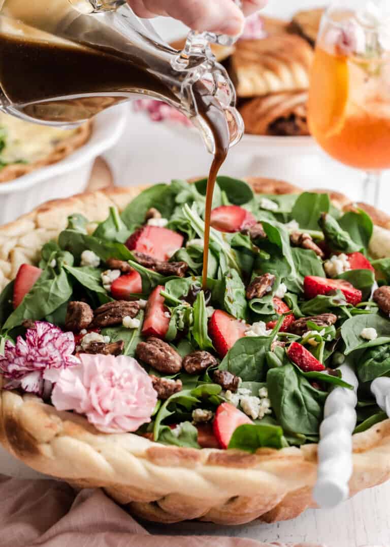 strawberry and spinach salad served in a bread basket bowl with cruet of vinaigrette dressing being poured on top, on white table.