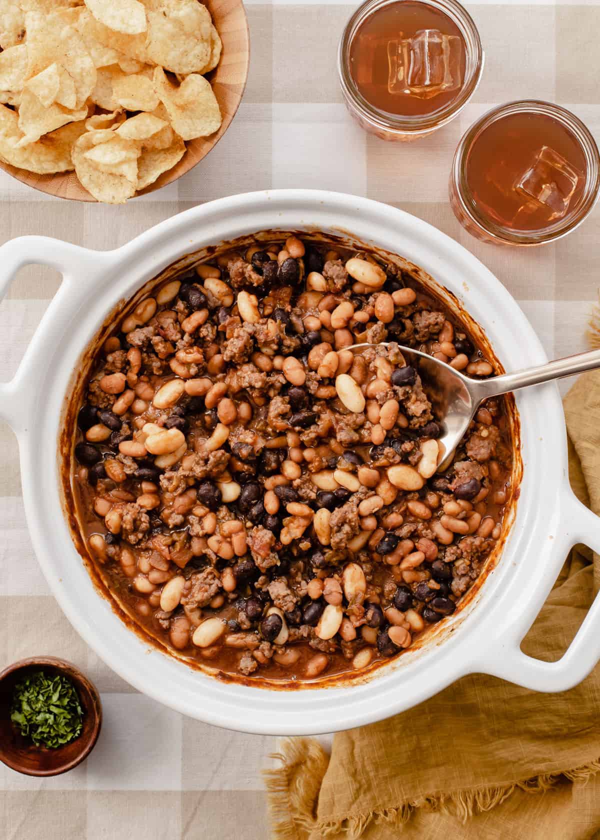 white round baking dish with baked beans surrounded by chips and drinks on table, overhead.