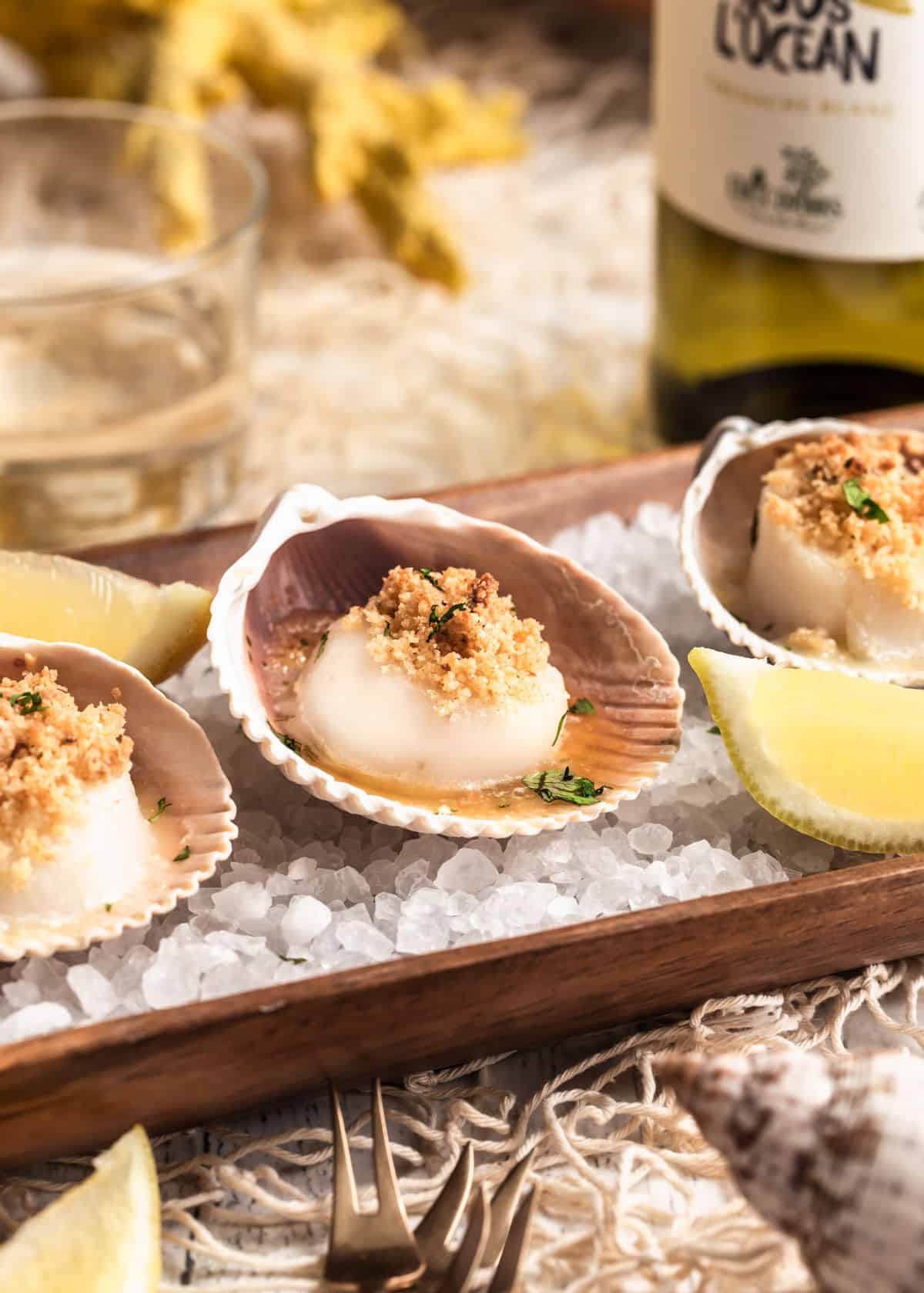close up of baked scallops topped with breadcrumbs, in shells on a bed of rock salt in wood serving tray.