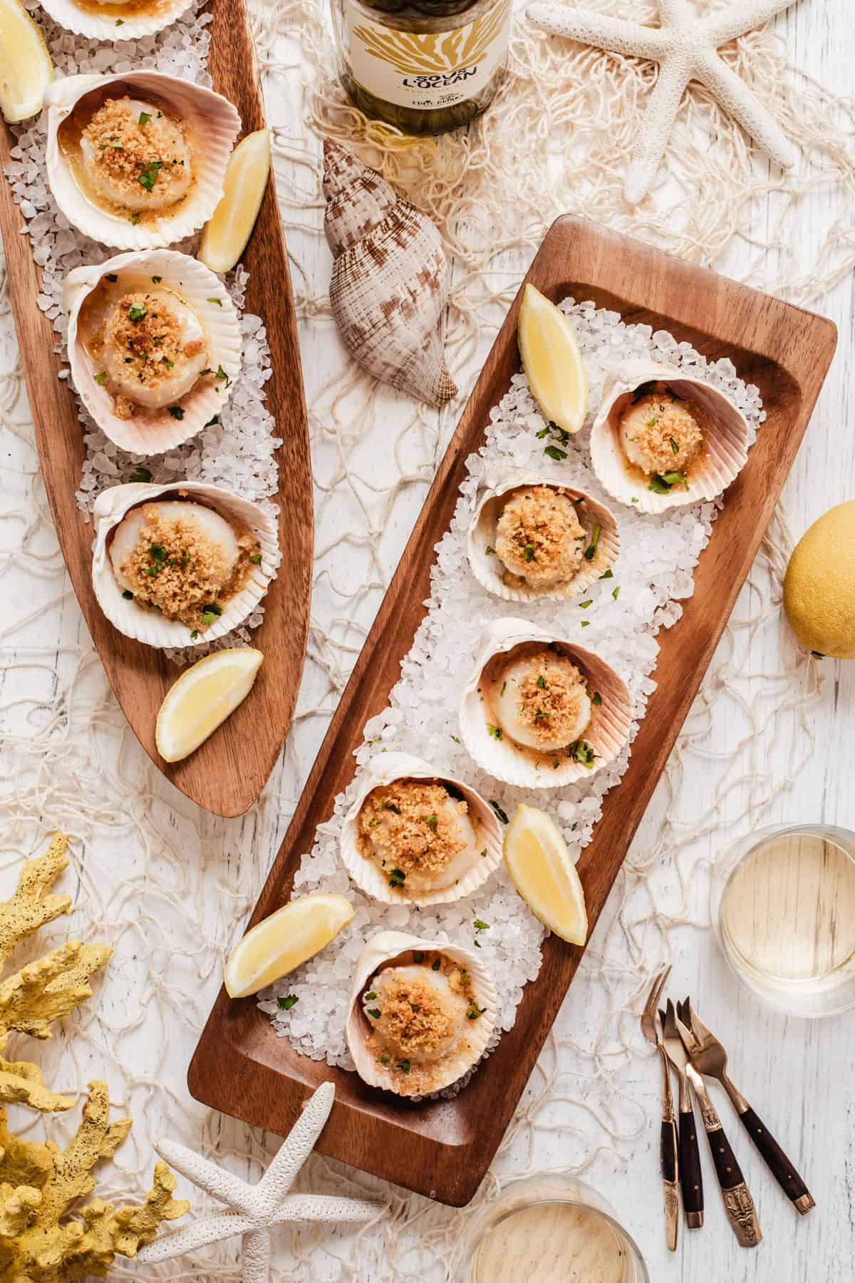 overhead view of long wood trays topped with scallops in shells on a bed of rock salt, surrounded by shells, fishnet, and wine.