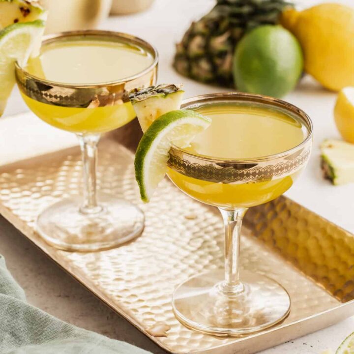 two coupe glasses filled with golden colored drink with pineapple and lime garnish.