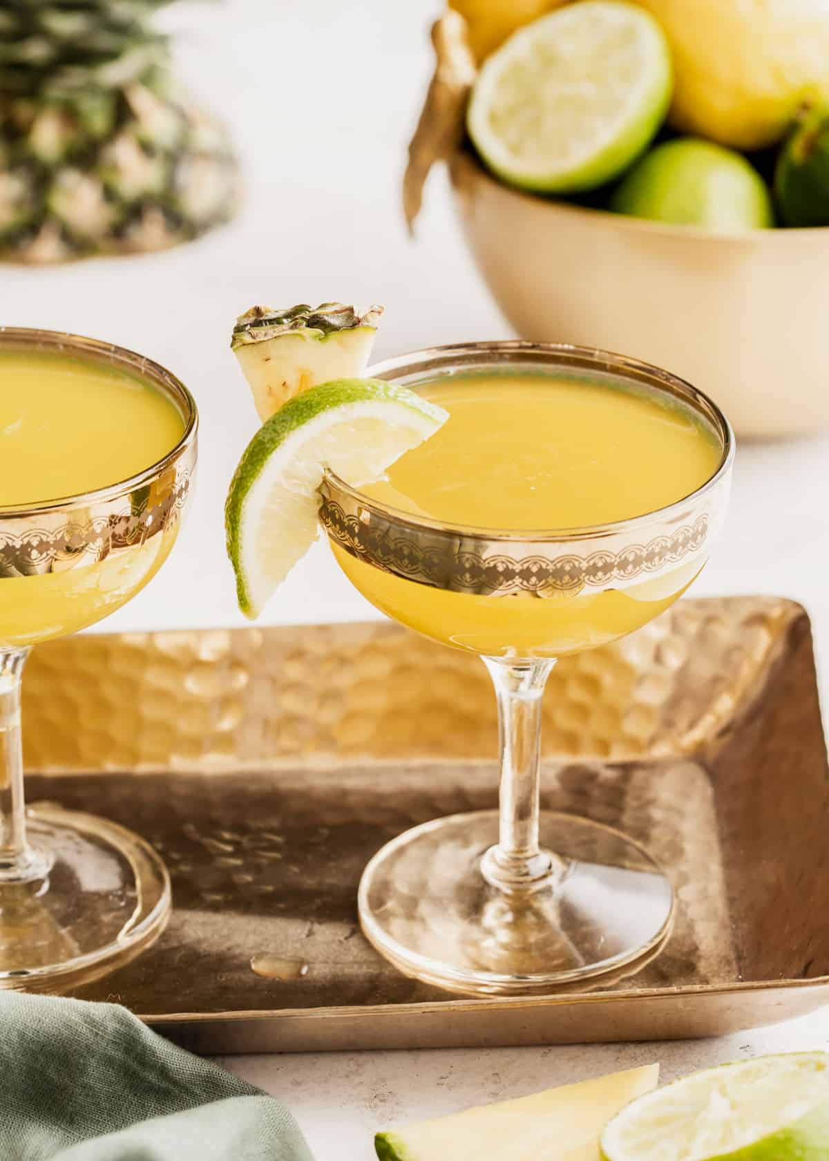 golden colored drink in coupe glass with lime and pineapple garnish, on gold tray.