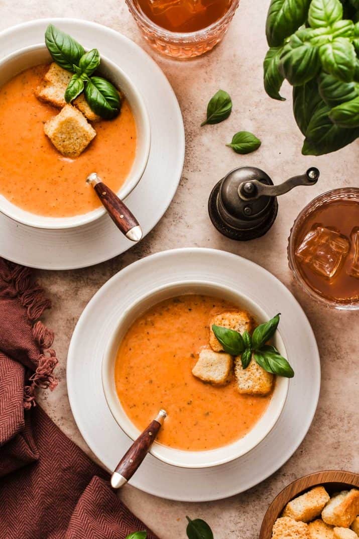 two bowls of tomato soup garnished with basil and croutons, on white plates with drinks at the side.