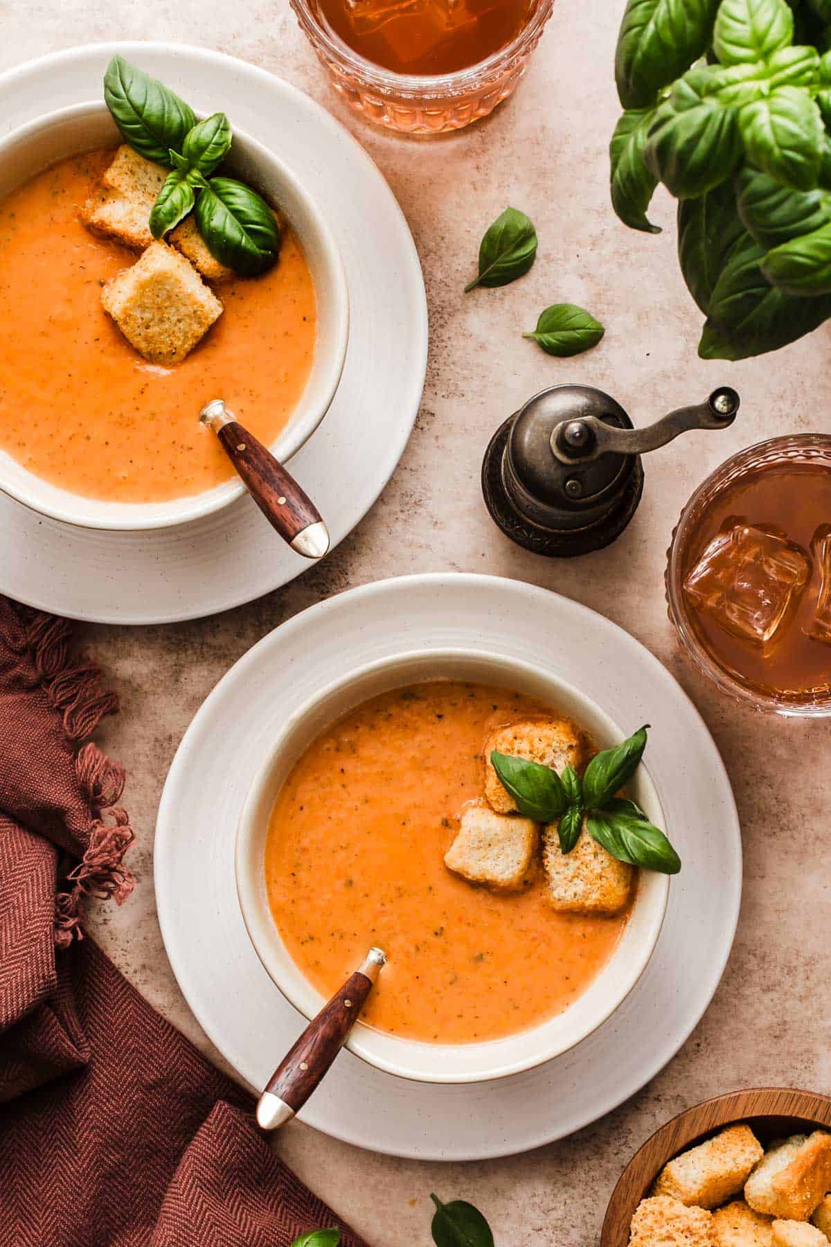 two bowls of tomato soup garnished with basil and croutons, on white plates with drinks at the side.