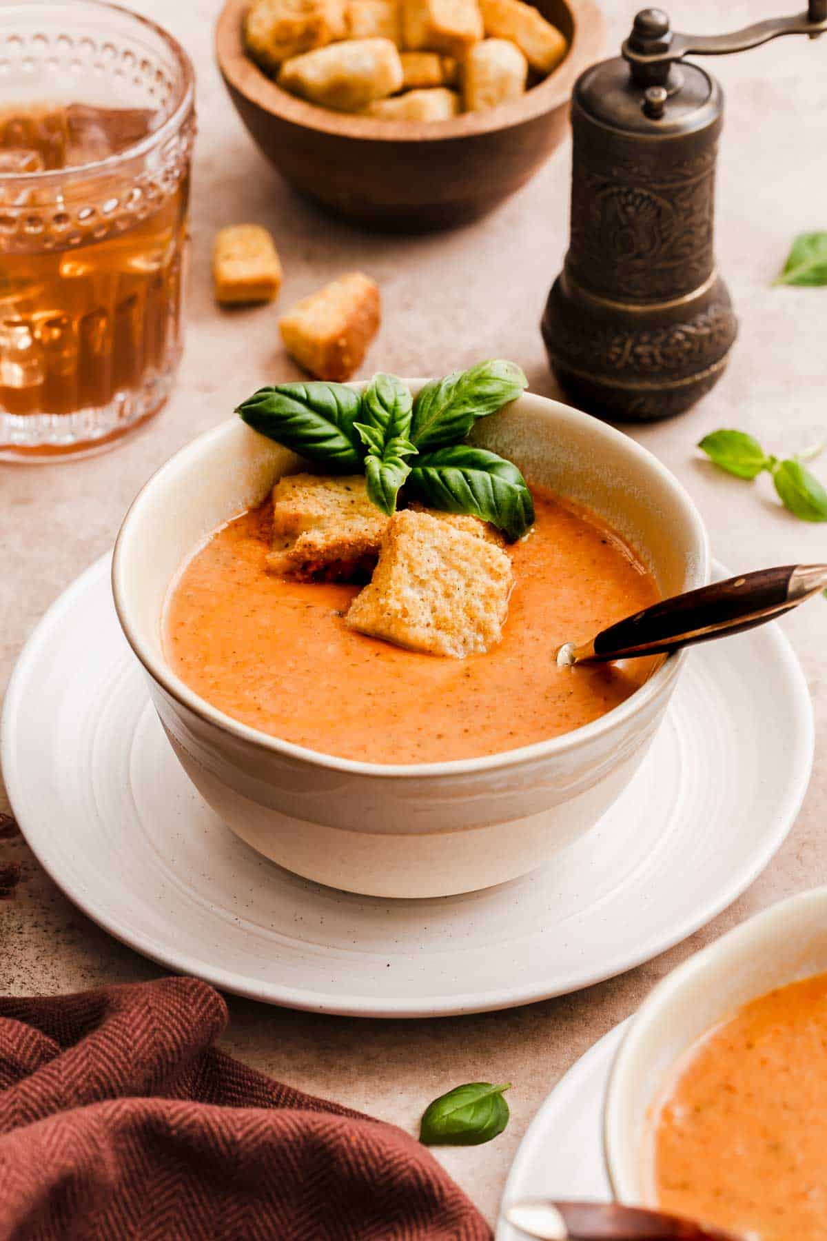 bowl of tomato soup garnished with fresh basil and croutons, on table with drink, napkin and pepper mill.