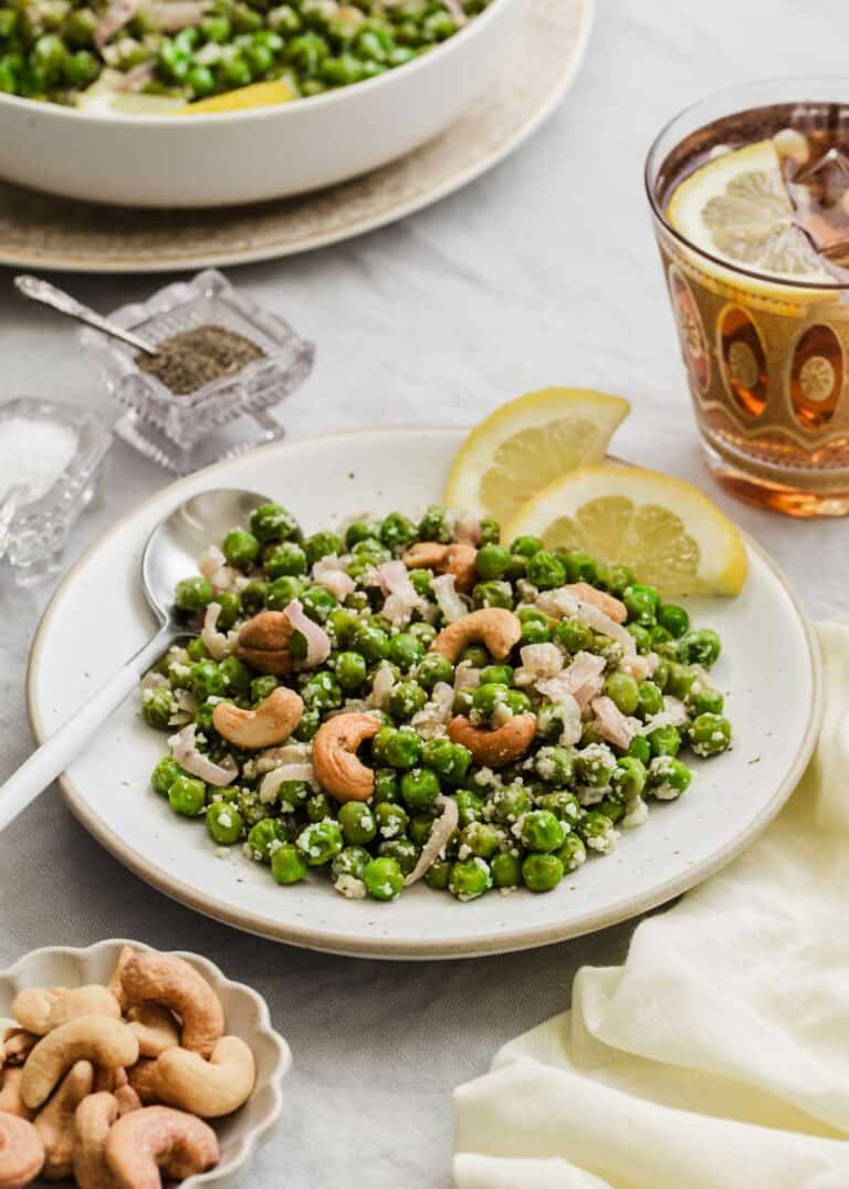 green peas topped with cashews on white plate with lemon wheel garnish, on table.