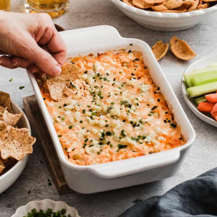 buffalo chicken dip in white baking dish with hand scooping a tortilla chip into it.