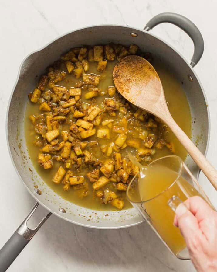 cooked chopped vegetables in large skillet with hand pouring in broth.