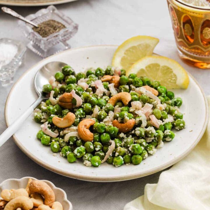 green peas topped with cashews on white plate with lemon wheel garnish, on table.