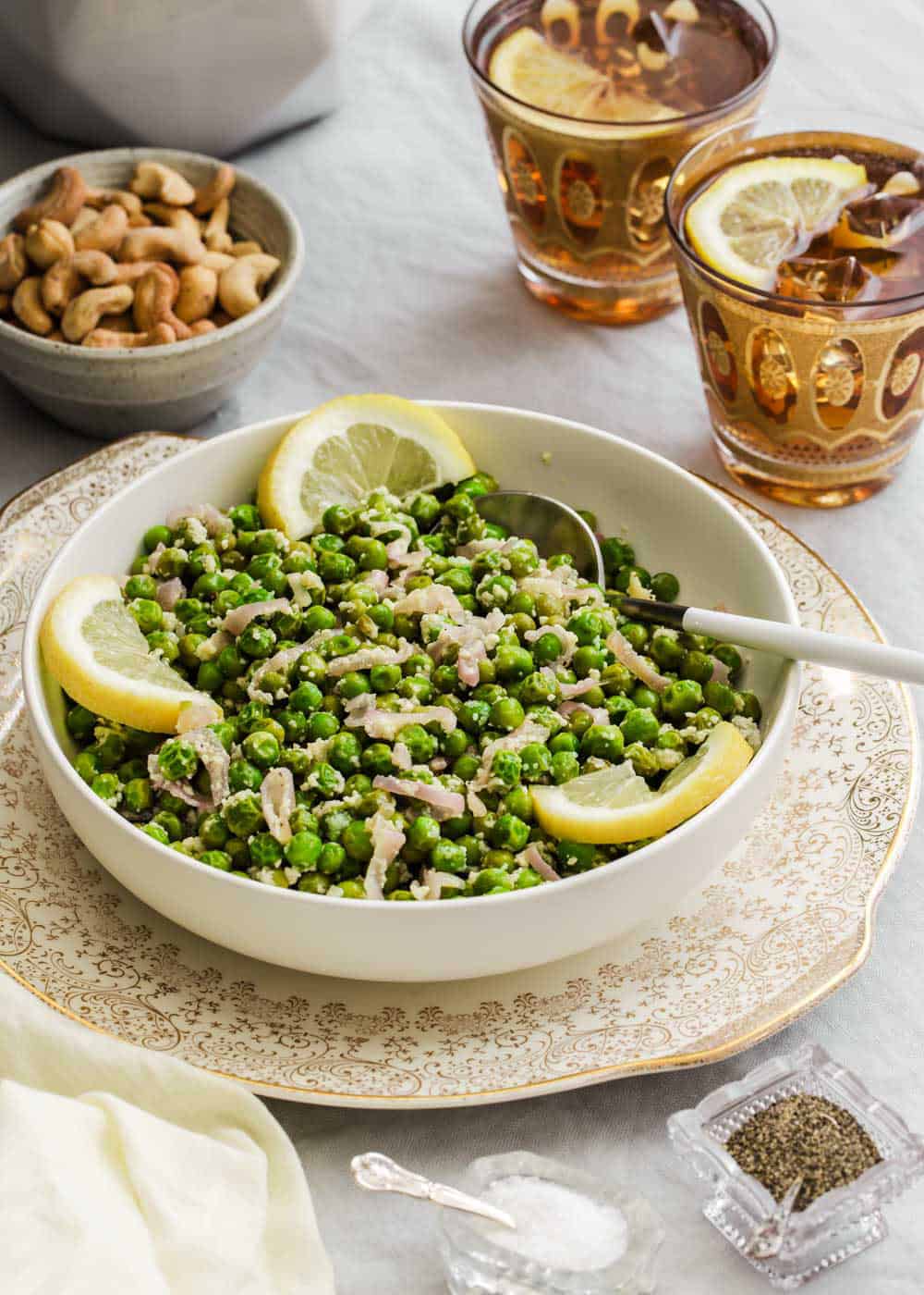 green peas in serving bowl with lemon wheel garnish, drink glasses and bowl of cashews on the side.