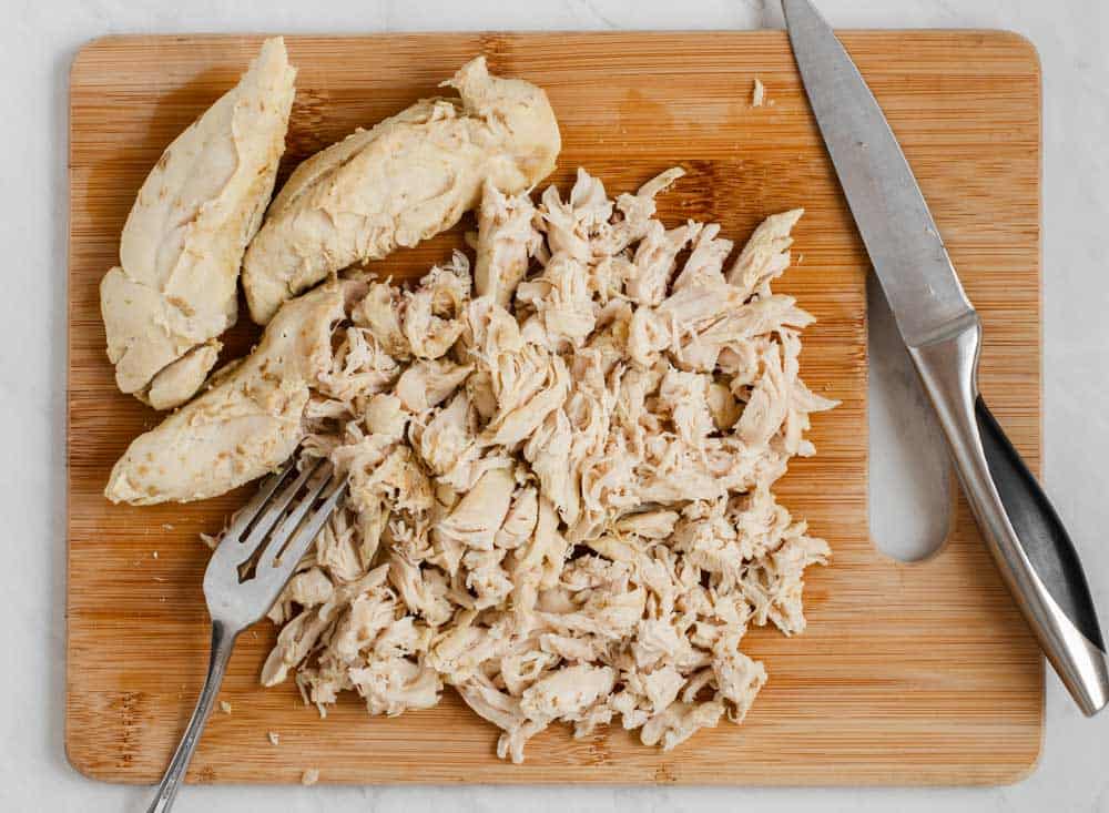 wood board with chicken breasts being shredded with fork and knife.