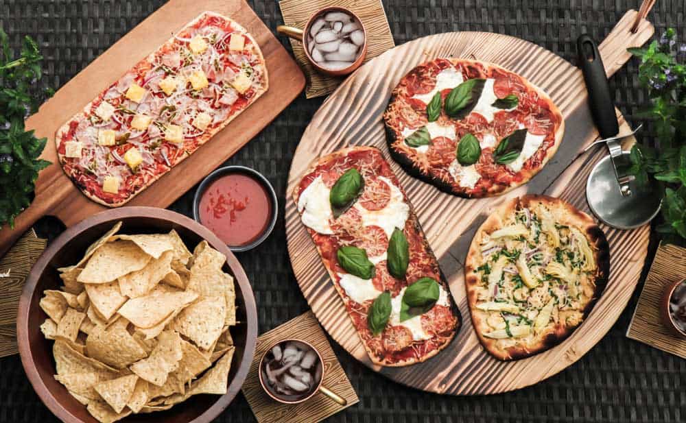 table with a variety of pizzas, along with chips and salsa, and drinks.